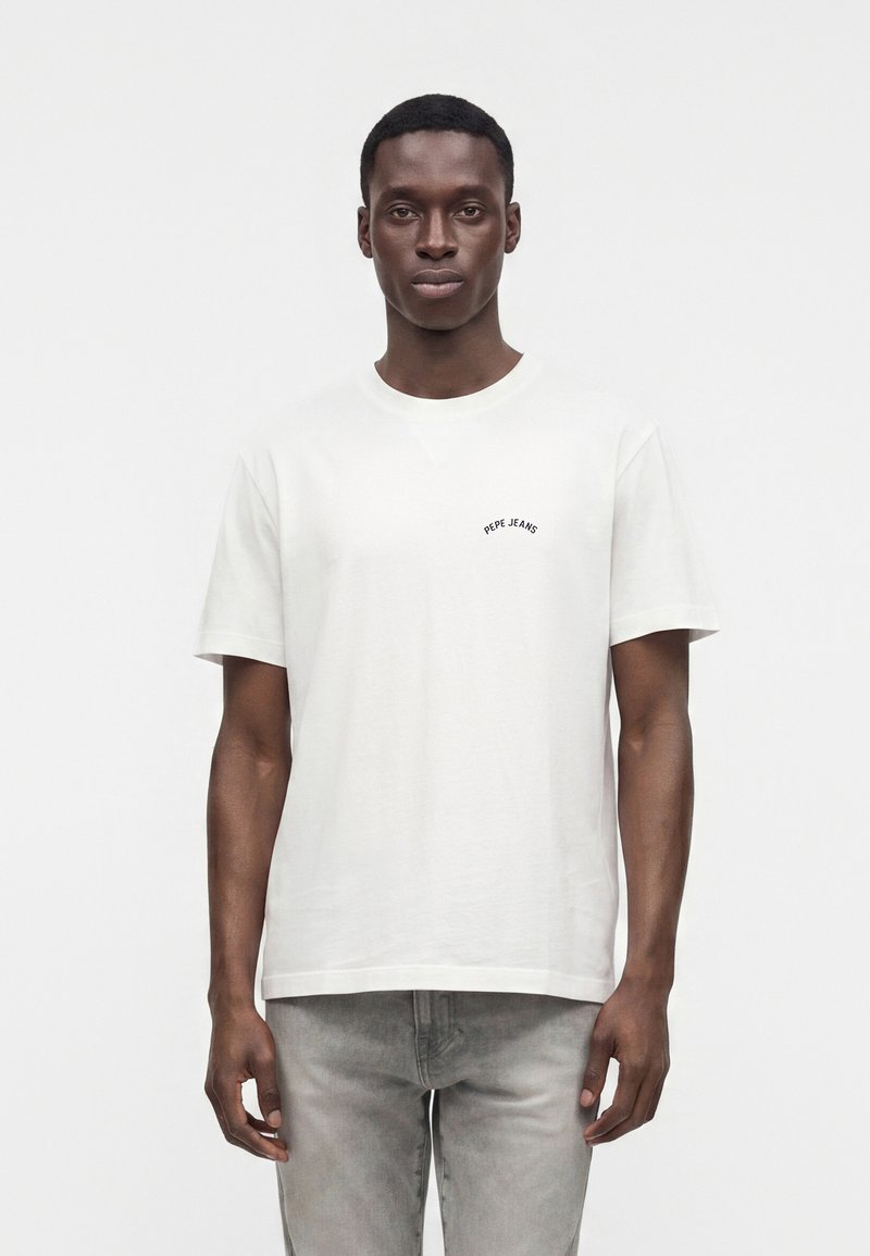 Άνδρας που φοράει ένα απλό λευκό T-shirt Pepe Jeans και ανοιχτό γκρι τζιν, στέκεται μπροστά σε λευκό φόντο, κοιτάζοντας προς τα εμπρός.