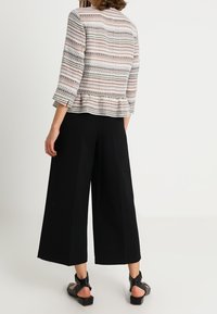 Knielange peplum top met meerkleurig gestreept kantpatroon, gecombineerd met zwarte wijde broek en platte zwarte sandalen met bandjes.