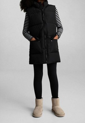 Gilet imbottito nero senza maniche con chiusura a zip e due tasche frontali, indossato sopra una camicia a maniche lunghe a righe bianche e nere e leggings neri, abbinato a stivali beige.