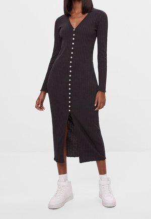 Femme portant une longue robe noire côtelée avec des boutons sur le devant, manches longues et baskets blanches montantes, debout devant un fond uni.