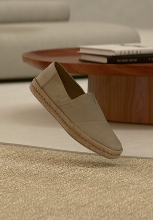 Scarpa beige senza lacci con suola intrecciata sospesa sopra un tappeto a trama, con un tavolo rotondo in legno e libri impilati sullo sfondo.