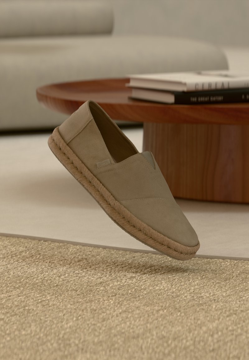 Chaussure beige à enfiler avec semelle tressée survolant un tapis texturé, avec une table ronde en bois et des livres empilés en arrière-plan.