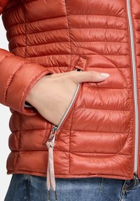 Rød quiltet pufferjakke med en lynlåslomme og et dekorativt lynlåstræk. Har en glat, skinnende overflade og en tætsiddende pasform.