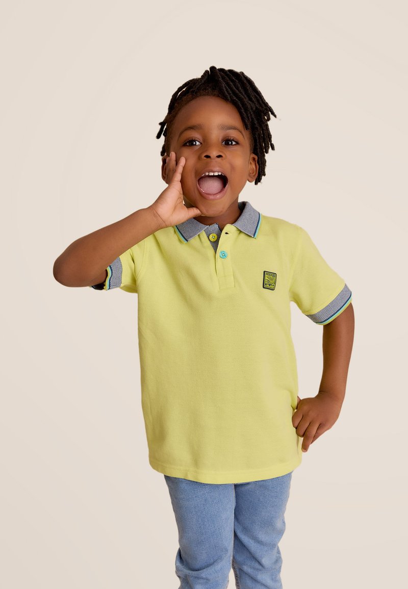 Lichtgele polo shirt met grijze kraag en manchetten, voorzien van blauwe knoopaccenten en een logo patch. G gedragen met blauwe jeans.