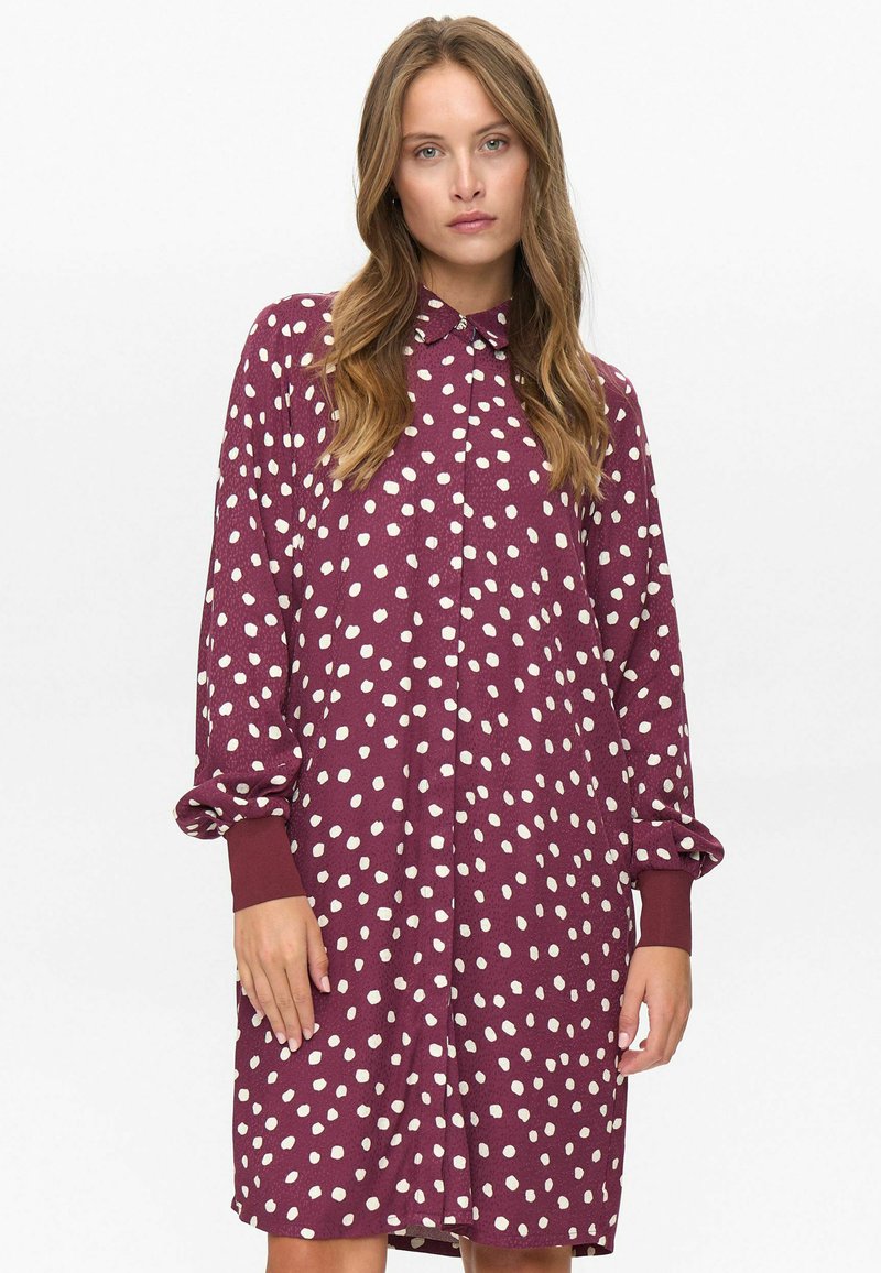 Femme portant une robe maroon à manches longues, à boutons, avec des pois blancs et des manches retroussées, se tenant devant un fond blanc uni.