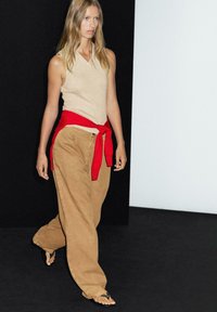 Top beige senza maniche, pantaloni khaki larghi, maglione rosso legato in vita, sfondo nero, modella che cammina, calzature casual con anelli per le dita.
