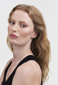 Donna con pelle chiara e lentiggini, capelli castano chiaro ondulati, che indossa un top nero, orecchini a mezzaluna dorati e una collana dorata abbinata.