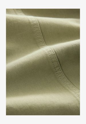 Hele oliivroheline kangas, millel on sile tekstuur, nähtavad õmblused ja kerge läikega, edastab pehme ja vastupidava välimuse.