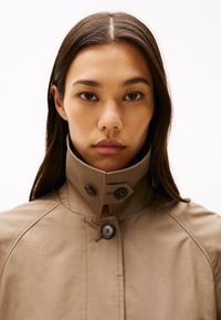 Giacca beige con colletto alto e una superficie liscia, caratterizzata da due bottoni scuri e leggeri dettagli di cucitura.