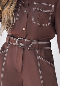 Mono marrón con costuras blancas en contraste, dos bolsillos en el pecho, una cintura ceñida con cinturón y una textura de tela suave, que exhibe un diseño moderno.