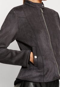 Giacca in finta pelle scamosciata nera con colletto alto, chiusura frontale con zip e pannelli perforati. Presenta bottoni a pressione argentati e texture a contrasto.
