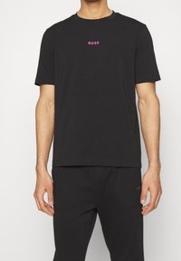 Svart kortärmad t-shirt i bomull med rund halsringning och en rosa "BOSS"-logotyp på bröstet. Slät textur och avslappnad passform.