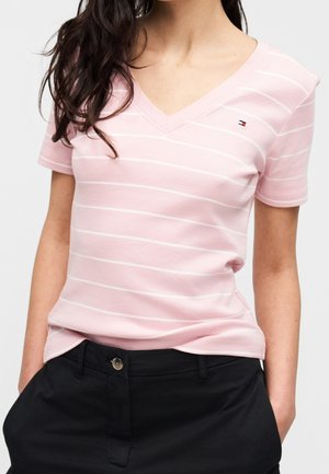 Frau trägt ein hellrosa Kurzarm-V-Ausschnitt-Shirt mit dünnen weißen horizontalen Streifen und schwarze Hosen mit Händen in den Taschen.