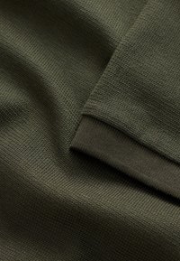 Maslinasto zelena tkanina s teksturiranom, uzorkovanom površinom s rebrastim rubom manžete. Mekani i gusto tkan materijal.