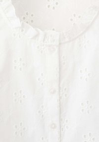 Blouse blanche avec une petite broderie ajourée en forme de fleur, col volanté et une rangée de boutons sur le devant.