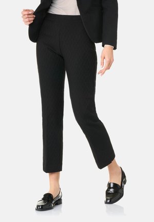 Frau trägt schwarze, strukturierte, schmal geschnittene Hose, schwarzen Blazer, weißes Oberteil und schwarze, glänzende Loafers, steht vor weißem Hintergrund.