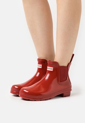 Bottines en caoutchouc rouge avec panneaux latéraux élastiques, une finition lisse et brillante, et des accents de logo à l'avant. Talon bas et semelle texturée.