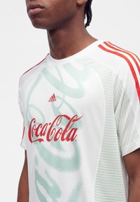 Άτομο που φοράει λευκό αθλητικό μπλουζάκι Adidas με κόκκινο λογότυπο Coca-Cola και τρεις κόκκινες ρίγες στον ώμο.