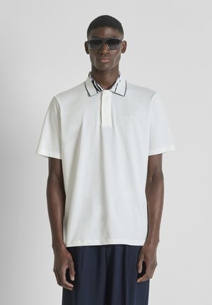 Modelo con polo blanco de manga corta con cuello estampado en blanco y negro y gafas de sol oscuras, de pie frente a un fondo gris liso.