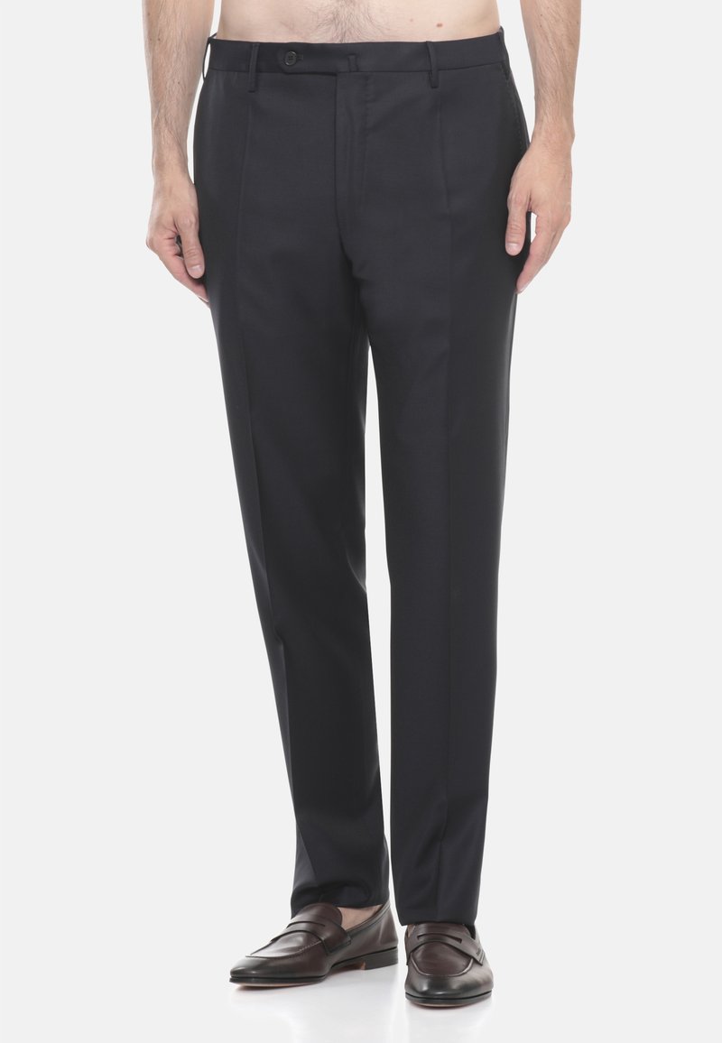 Uomo che indossa pantaloni eleganti blu navy su misura e mocassini in pelle marrone, in piedi contro uno sfondo chiaro e semplice.