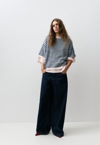 Ung kvinde med langt hår iført en løs stribet sweater og brede mørke jeans, hænderne i lommerne, stående mod en ensfarvet lys baggrund.