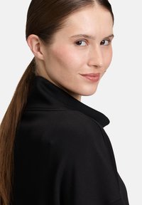 Schwarzer, weicher Stoffmantel mit Kragen, der eine glatte Textur und ein strukturiertes Design aufweist, getragen von einer Frau mit langem, zurückgebundenem Haar.