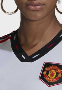 Camiseta blanca del Manchester United con un cuello en V negro, que presenta acentos de costura en rojo y blanco y un escudo del club bordado.
