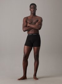 Schwarze Baumwollboxershorts mit taillierter Passform und breitem elastischem Bund mit dem "Calvin Klein" Logo. Getragen von einem stehenden männlichen Model.