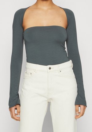 Femme portant un haut gris foncé à manches longues et épaules dénudées associé à un jean blanc taille haute, debout devant un fond uni.