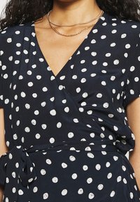 Námořnické zavinovací šaty s bílými puntíky, krátkým rukávem a detailem vázání v pase. Hladká látka s jemným leskem.