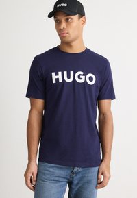 Jūras zila kokvilnas T krekls ar baltu "HUGO" tekstu, apa�ļa kakla, īsām piedurknēm, kombinēts ar melnu cepuri ar to pašu logo baltā krāsā.