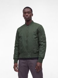 Homme portant une veste bomber zippée vert foncé et un pantalon sombre, debout devant un fond blanc uni.