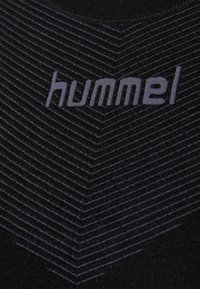 Logo Hummel en gris sur tissu noir avec de fines lignes chevrons grises formant un motif en V sur la surface textile.