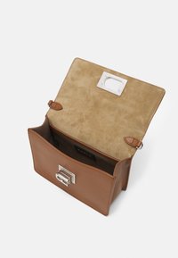 Brązowa skórzana torba z klapką, ukazująca beżowe, zamszowe wnętrze. Posiada srebrną klamrę, minimalistyczny design i wewnętrzne logo.