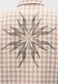 Μπεζ καρό πουκάμισο με ένα μεγάλο, σκούρο γραφικό σχέδιο που μοιάζει με φλόγες στην πλάτη. Το ύφασμα φαίνεται υφασμένο με υφή και σχέδιο ύφανσης.