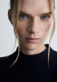 Ritratto di una persona con capelli biondo chiaro e occhi blu, che indossa un maglione nero. La pelle ha una texture liscia con nei naturali.