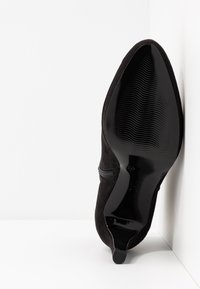 Botte en daim noir à bout pointu avec semelle texturée brillante et petit talon, positionnée verticalement contre un mur d'angle blanc.