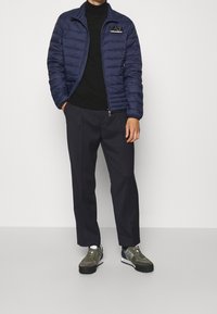 Marinblå pufferjacka med dragkedja och ståkrage, svart polotröja, mörka byxor och gröna sneakers med svarta detaljer.