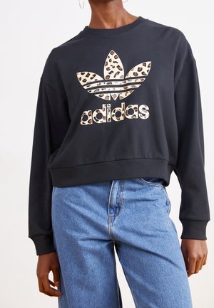 Person trägt schwarzen Adidas-Sweatshirt mit Leopardmuster-Logo und blaue hoch taillierte Jeans, steht vor weißem Hintergrund.