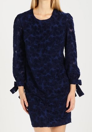 Dunkelblaues Kleid aus floralem, strukturiertem Stoff, mit langen, transparenten Ärmeln und Schleifenakzenten an den Manschetten. Rundhalsausschnitt und figurbetonter Schnitt.