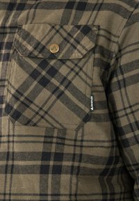 Bruin en zwarte geruite flanellen blouse met een voorkeurszak met een messing knoop en een logotag aan de zijkant. Zachte textuur en duurzaam materiaal.
