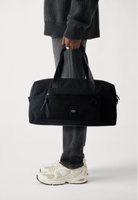Schwarze Sporttasche mit einer Reißverschluss-Vordertasche, stabilen Griffen und einem Logo-Patch. Glattes Material mit minimalistischem Design.