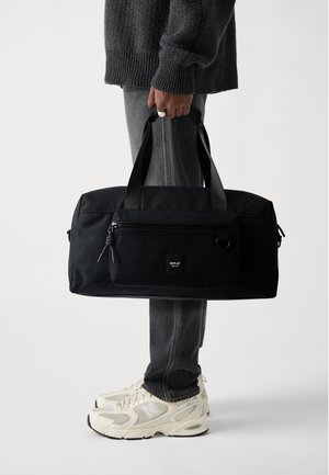 Bolsa de deporte negra con un bolsillo frontal con cremallera, asas resistentes y una parche con logo. Tejido suave con un diseño minimalista.