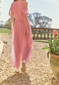 Robe longue rose avec un design épaule dénudée, tissu doux et silhouette fluide ; associée à des chaussures blanches, dans un jardin extérieur.