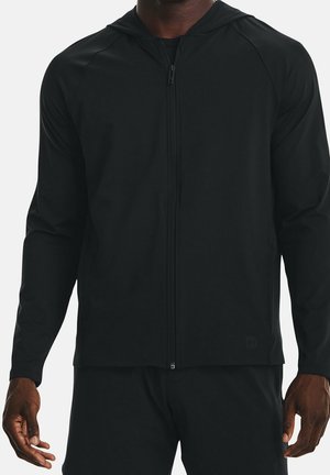 Mand iført en ensfarvet sort lynlåstrøje og matchende sorte bukser, vist fra skuldre til hofter mod en ensfarvet baggrund.