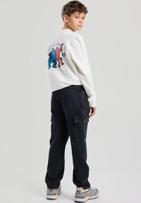 Felpa bianca con grafica blu e rossa sul retro; pantaloni cargo neri con tasche; sneakers grigie. Il modello posa di lato.