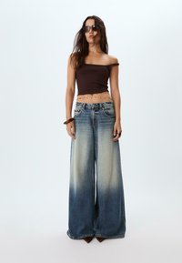 Smeđi crop top s otkrivanim ramenima, svijetloplave široke traperice s gradijentnim izblijedjenjem, dodatno ukrašen slojevitim lančićem i velikim crnim sunčanim naočalama.