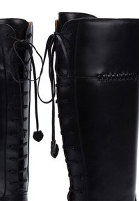 Las botas altas de cuero negro presentan un diseño elegante, cordones en la parte frontal y acentos trenzados a lo largo de la costura. Textura suave.