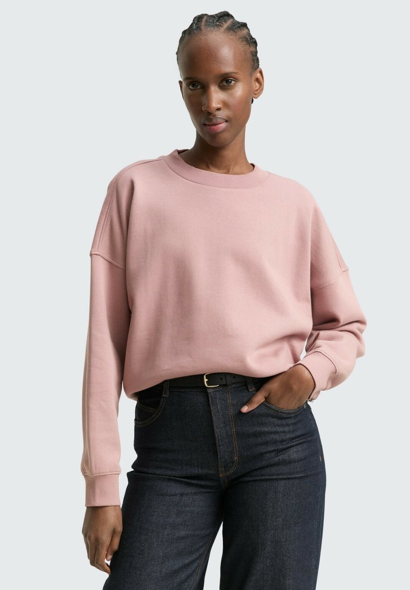 Sudadera rosa claro con cuello redondo y hombros caídos, combinada con vaqueros de denim azul oscuro. Material suave y corte relajado.