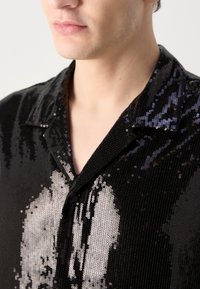 Homme portant une chemise noire à sequins avec col, montrant un motif argenté réfléchissant à l'avant.